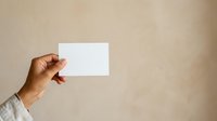 Male hand with medium skin tone holding blank white business card against neutral beige wall background for corporate branding mockup and professional identity presentation.