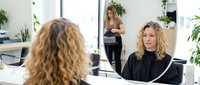 Blonde woman getting professional hair styling treatment at modern salon with stylist working in background surrounded by green plants.