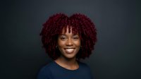 Young woman with deep burgundy curly hair, warm brown skin tone, and bright smile wearing dark navy top against neutral background for portrait use.