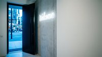 Glowing neon welcome sign on concrete wall beside open door leading to busy city street, modern interior entrance with urban backdrop.
