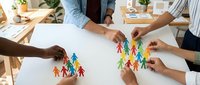 Diverse group of people arranging colorful paper cutout figures on white surface, representing teamwork, community building, and collaborative planning for business or organizational projects.