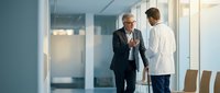 Senior male executive in dark suit and glasses conversing with younger male colleague in white shirt in modern office hallway with glass partitions, suitable for corporate business communications.