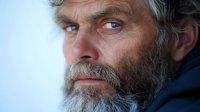 Dramatic close-up portrait of mature bearded man with intense blue eyes and gray hair looking thoughtfully into distance against soft light background.