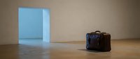 Vintage leather travel bag sits on wooden floor beside glowing blue doorway portal in minimalist room with warm beige walls creating surreal atmosphere.