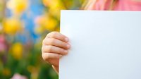 Child's hand holding blank white card with colorful flower garden background, suitable for greeting cards, announcements, and promotional messaging.