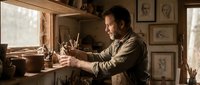 Focused Caucasian male craftsman sculpting a figurine in a rustic wooden workshop filled with tools, sketches, and warm natural light.