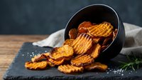Crispy golden potato chips spilling from black bowl with sea salt and fresh herbs on rustic wooden table for snack food and cooking concepts.