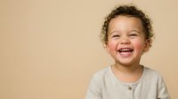 Toddler with curly hair and mixed ethnicity smiling brightly against neutral beige background, suitable for parenting, childcare, and family-related designs.