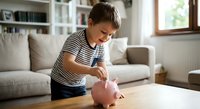Young boy in striped shirt putting coin into pink piggy bank at home, teaching children about saving money and financial literacy for future goals.