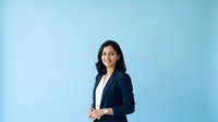 Professional woman with dark hair and South Asian ethnicity wearing navy blazer over white shirt, standing confidently against light blue background. Suitable for corporate, business, and professional website use.