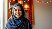 Happy young Middle Eastern woman wearing blue hijab smiling warmly at camera with festive holiday lights and decorations creating cozy atmosphere in background.