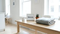 Minimalist bright interior with wooden table, welcome sign, folded blanket, mug, and small plant by sunlit windows.