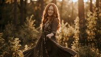Young woman with long curly red hair in flowing black dress dancing in golden sunlight among autumn foliage creating magical atmospheric portrait.
