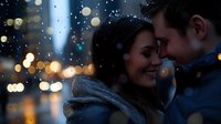 Young romantic couple embracing outdoors on winter evening with falling snow and blurred city lights bokeh creating magical atmosphere for Valentine cards.