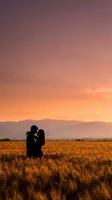 Romantic couple silhouette embracing in golden wheat field at sunset with mountains and colorful sky for wedding and love concepts.
