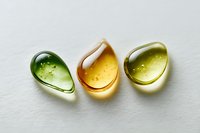 Three colorful glass droplets with bubbles in green, amber and yellow tones arranged on white surface for wellness and beauty product design concepts.