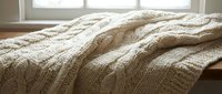 Cozy cream cable knit blanket draped over furniture near bright window creating warm comfortable home atmosphere for relaxation and interior design.