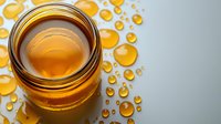 Golden honey in glass jar with scattered drops on white surface, macro food photography showing natural sweetener texture and amber transparency.