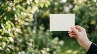 Close-up of hand holding blank white card against blurred green foliage background, suitable for business cards, invitations, and marketing mockups.