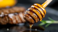 Honey dripping from wooden dipper onto grilled meat, close-up macro shot showing golden syrup texture and glossy surface against dark background.