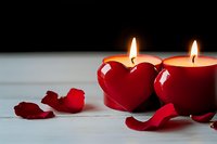 Two red candles glowing warmly beside a shiny heart with rose petals on white wood, evoking romance and love for Valentine's Day.
