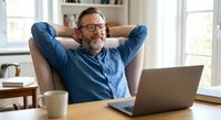 Relaxed middle-aged Caucasian businessman with glasses and beard sitting in armchair with hands behind head, smiling while working on laptop at home office.