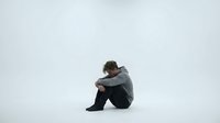 Young man sitting alone in contemplative pose against minimalist white background expressing solitude, depression, mental health struggles and emotional isolation.