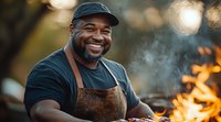African American male chef in leather apron smiling while grilling outdoors at sunset, steam rising from barbecue with fire and bokeh lights in background creating warm atmosphere.