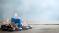 Blue candle burning with white lily flowers and smooth stones on wooden surface creating peaceful spa atmosphere for relaxation and wellness.