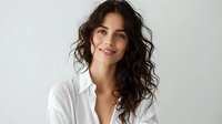 Young woman with long dark curly hair wearing white shirt smiling at camera against light background. Natural beauty portrait with minimal makeup and casual style.