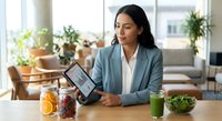 Young Hispanic businesswoman in blue blazer reviewing analytics on tablet at desk with healthy food, green smoothie, and fresh fruit in bright modern office.