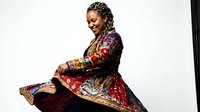 Pregnant woman with dark skin tone wearing ornate traditional patterned garment with gold embroidery, sitting in profile pose against white background for maternity and cultural fashion concepts.