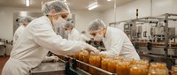 Food production workers in white uniforms and hairnets processing jars in commercial kitchen facility for quality control and packaging operations.