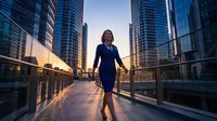Confident businesswoman in blue dress walking on modern city bridge at sunset with glass skyscrapers. Corporate success and urban lifestyle concept.