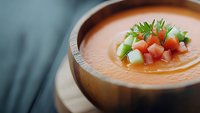 Refreshing gazpacho soup garnished with diced cucumber and tomatoes in wooden bowl, selective focus on fresh vegetable toppings and creamy orange texture.