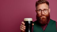 Bearded hipster man in glasses and green sweater holding dark craft beer with foam head against burgundy background. Professional portrait for brewery marketing.