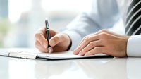 Caucasian business professional in white shirt signing document or writing notes on paper at office desk, close-up view of hands holding pen.