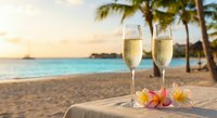 Two champagne flutes filled with sparkling wine on tropical beach at sunset with palm trees and turquoise ocean, romantic vacation celebration concept.