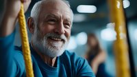 Happy senior Caucasian man with white beard smiling while exercising with resistance bands in modern fitness center for healthy aging and wellness.