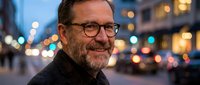 Middle-aged Caucasian businessman with glasses smiling confidently on urban street at dusk with bokeh city lights creating warm atmospheric background.