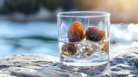 Glass of whiskey with ice cubes and dates on natural stone against blurred water background, golden hour lighting creates luxurious mood for beverage photography.