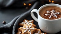 Hot chocolate with snowflake latte art pattern in white ceramic cup, served with decorated gingerbread star cookies on dark background with bokeh lights.