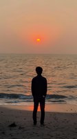 Silhouette of young man standing alone on beach watching orange sunset over ocean horizon, peaceful contemplative moment with waves and sandy shore.