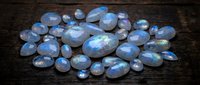 Collection of polished moonstone gems with blue flash adularescence scattered on dark wooden surface for jewelry making and crystal healing practices.