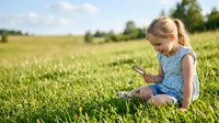 Young blonde girl in blue dress sitting on grass examining nature with magnifying glass in sunny meadow.