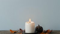 Lit white pillar candle surrounded by autumn leaves and dark decorative pumpkins on wooden surface against neutral background for fall seasonal decor and holiday styling.