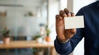 Man with dark skin tone wearing navy blazer holding blank white business card in modern office environment with plants and natural lighting.
