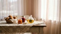 Cozy morning tea setup with glass teapot, jam jars and fresh bread in wicker basket on lace tablecloth by sunny window creating warm homey atmosphere.