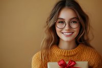 Young Caucasian woman with glasses and bright smile holding wrapped gift box with red bow, wearing cozy orange knit sweater against warm brown background.