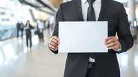 Professional businessman in dark suit holding blank white paper sign in modern office lobby with blurred people walking in background for corporate messaging.
